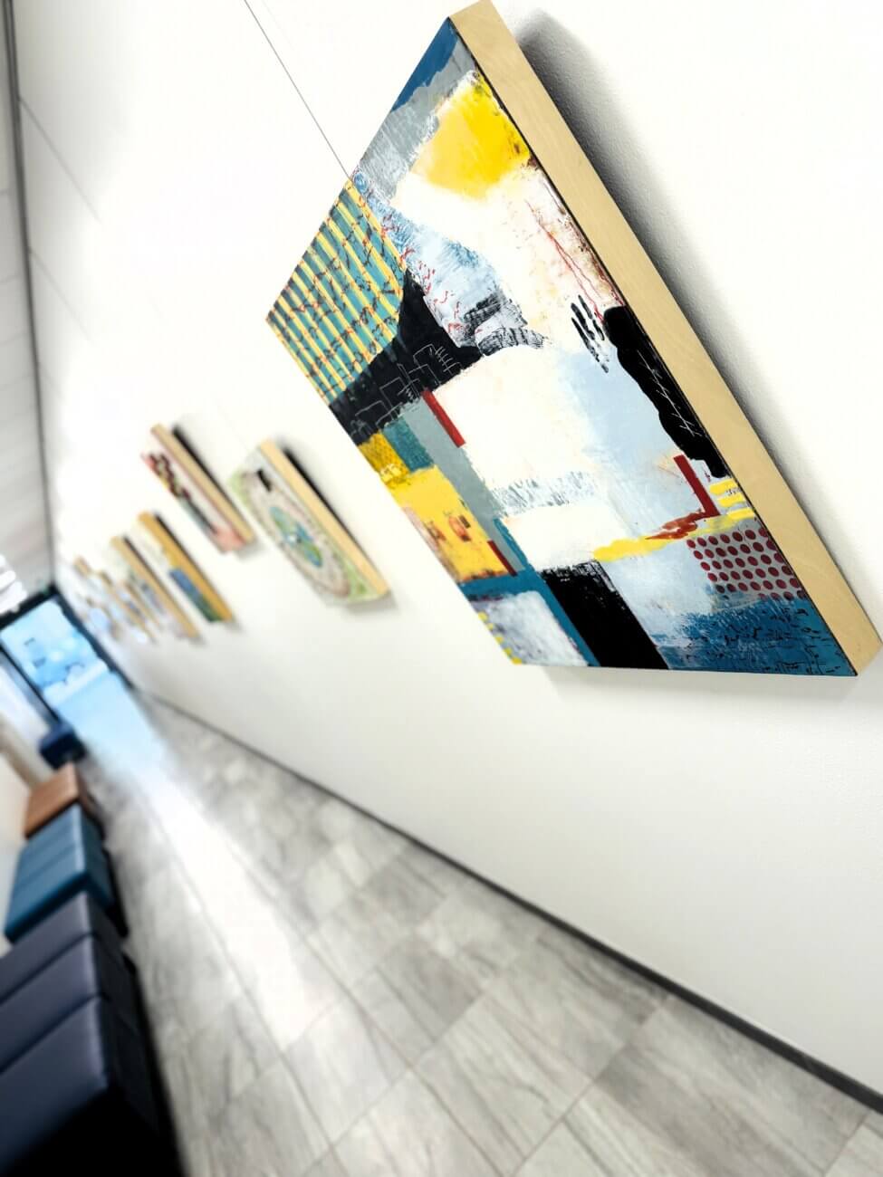 A close-up of abstract paintings in wooden frames hanging on a white wall along a bright, modern hallway at an art gallery, with marble floors and blue seating visible in the distance.
