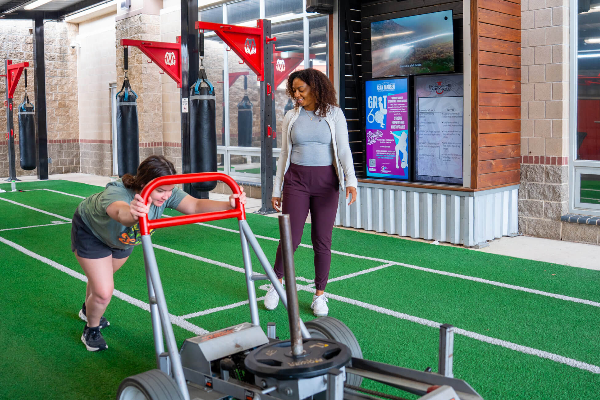 A woman pushes a weighted sled across indoor turf while another woman, likely a trainer, watches and encourages her in a gym with punching bags and digital displays in the background.