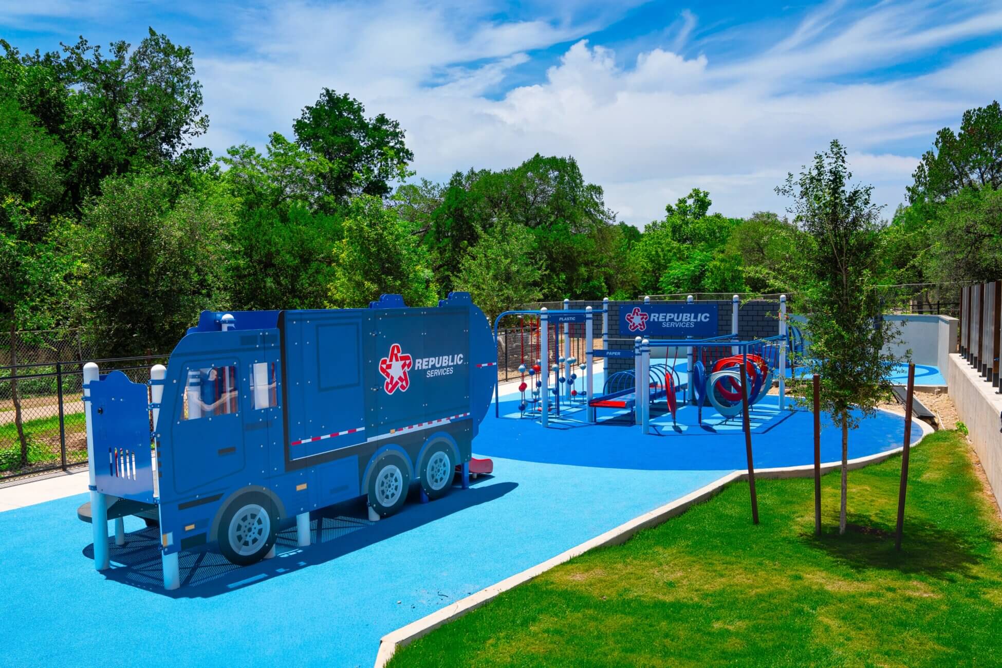 A playground with a large blue play structure shaped like a recycling truck and other climbing equipment sits on blue safety surfacing, surrounded by green trees and grass under a partly cloudy sky.