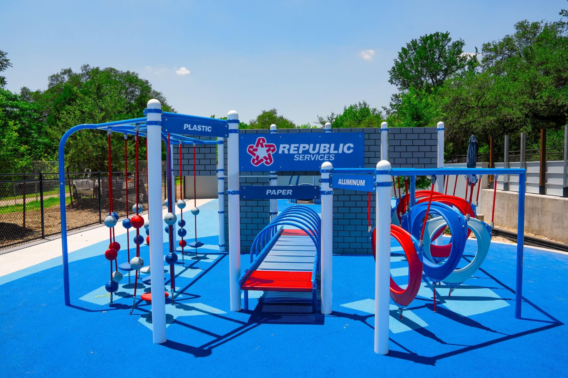 A colorful outdoor playground with recycling-themed play structures labeled “Plastic,” “Paper,” and “Aluminum” and a wall displaying the Republic Services logo, set on a bright blue rubber surface. Trees and a fence are in the background.