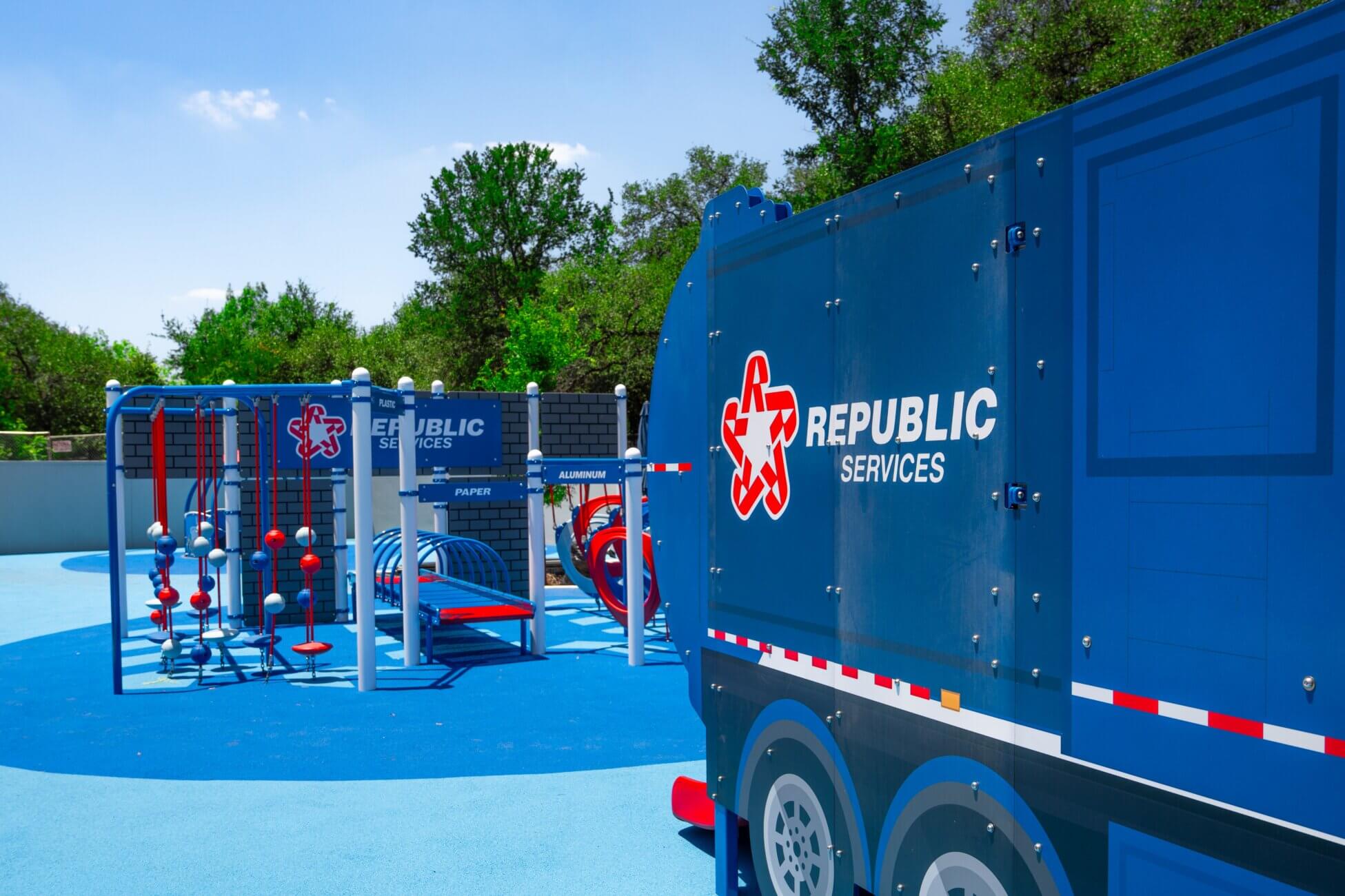 A playground with blue play structures and a large play truck, both branded with Republic Services logos, set on a bright blue surface with trees in the background.