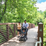 A family with a stroller walks together on a wooden bridge surrounded by lush green trees and foliage on a sunny day.