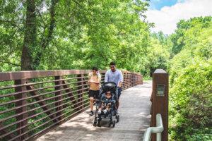 A family with a stroller walks together on a wooden bridge surrounded by lush green trees and foliage on a sunny day.