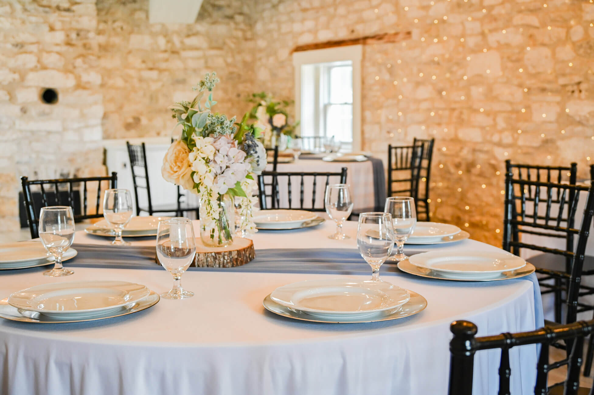 A round table with a white tablecloth is set with plates, glasses, and a floral centerpiece in a rustic room with stone walls, black chairs, and soft string lights in the background.
