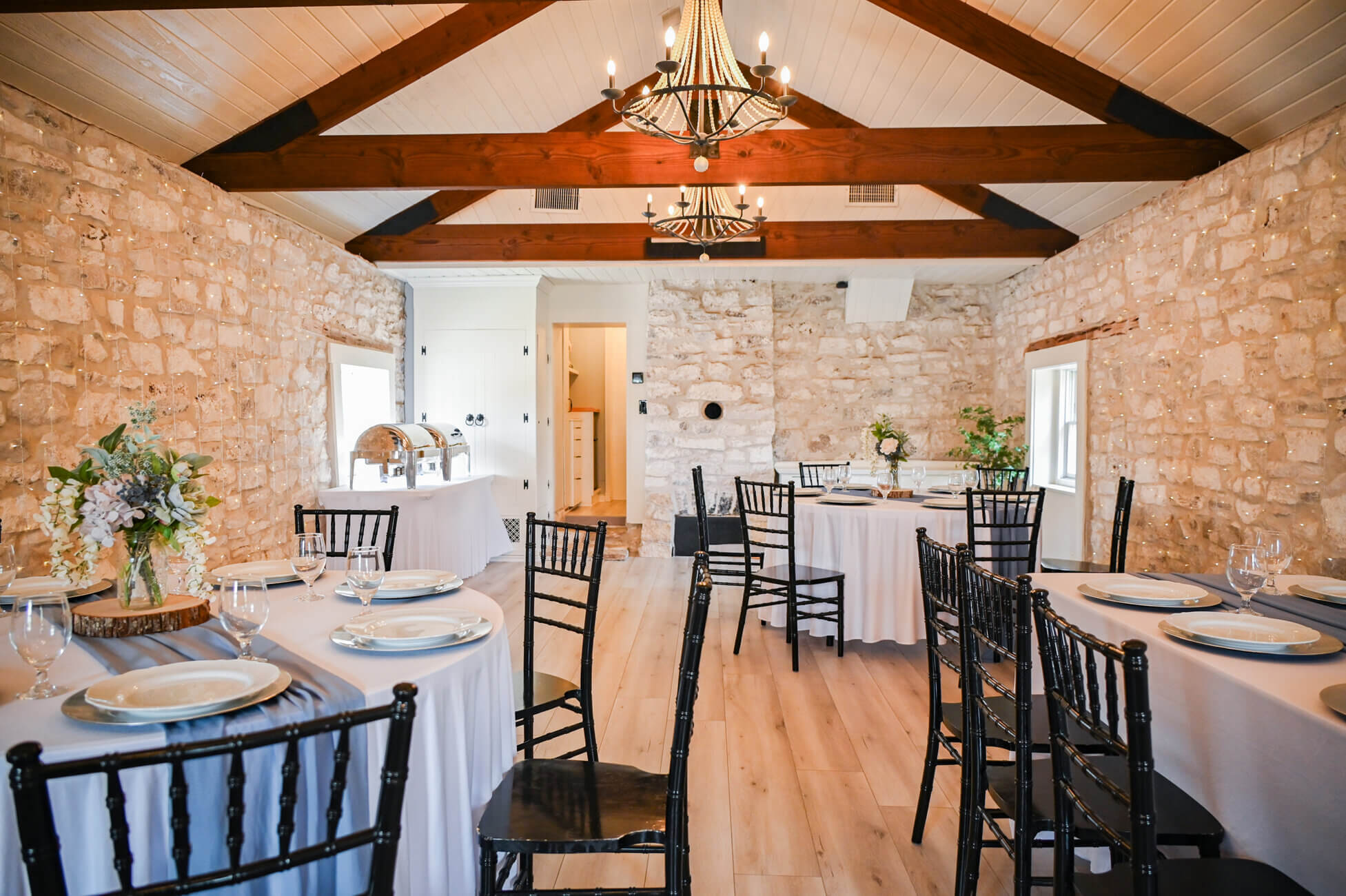 A rustic dining room with stone walls, wooden beams, round tables covered in white tablecloths, black chairs, floral centerpieces, and place settings, set for a meal under elegant chandeliers.