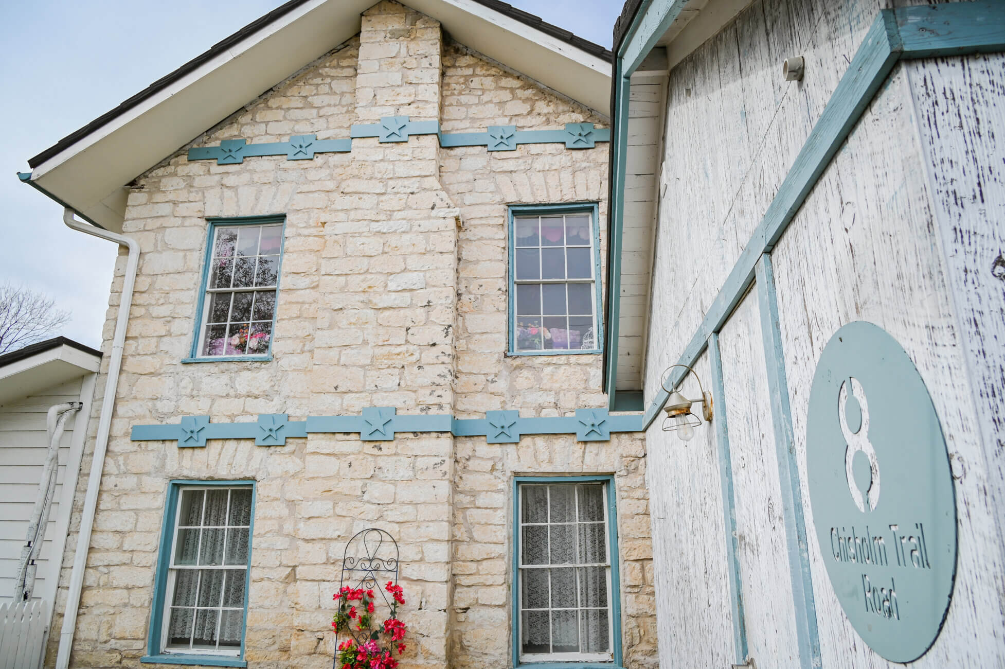 A stone house with blue trim and two windows with pink flowers. Beside it is a rustic white wooden building with a round blue sign reading 8 Chisholm Trail Road.