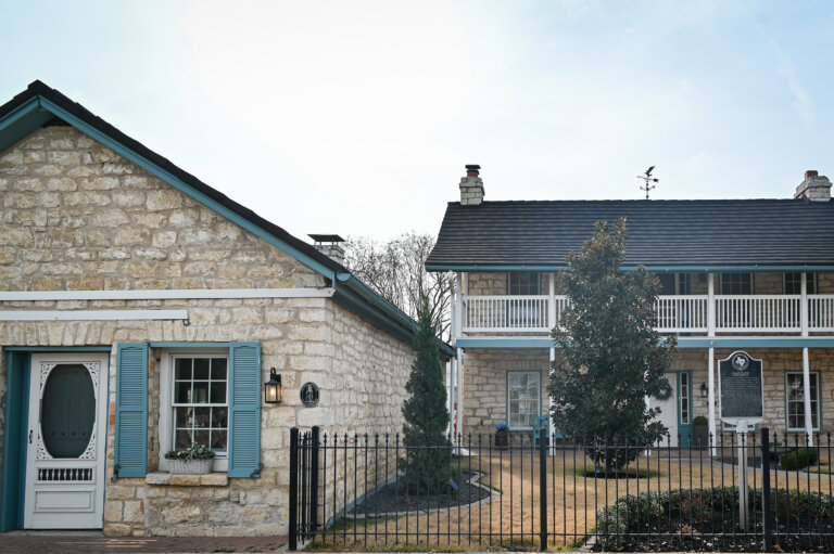 A historic stone building with blue shutters stands next to a two-story stone house with a balcony, white railings, and a black metal fence in front, on a sunny day.
