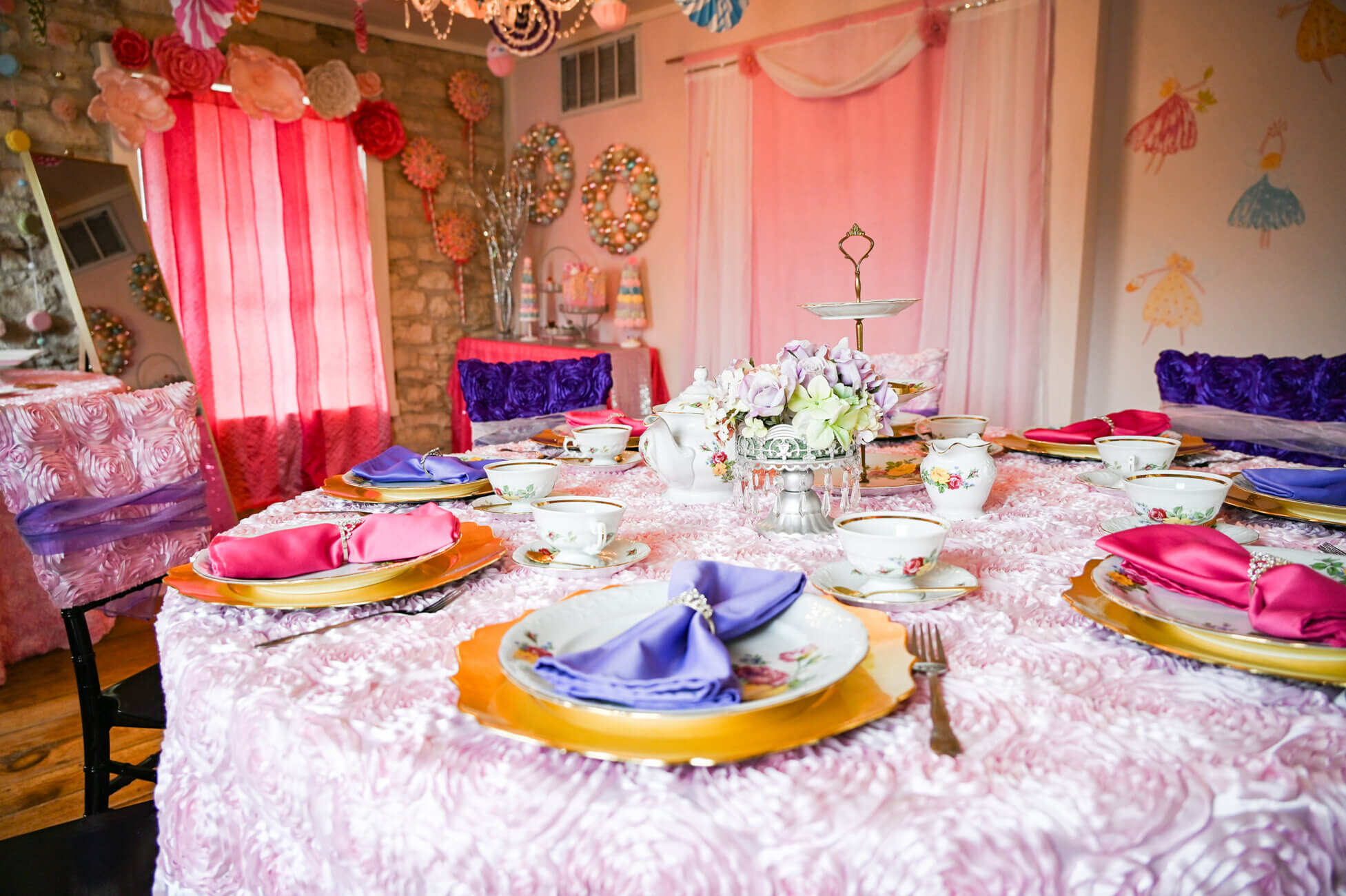 A round table set for a tea party with pastel-colored napkins, floral teacups, gold chargers, and a flower centerpiece. The room is decorated with pink and purple fabrics, flowers, and whimsical wall art.