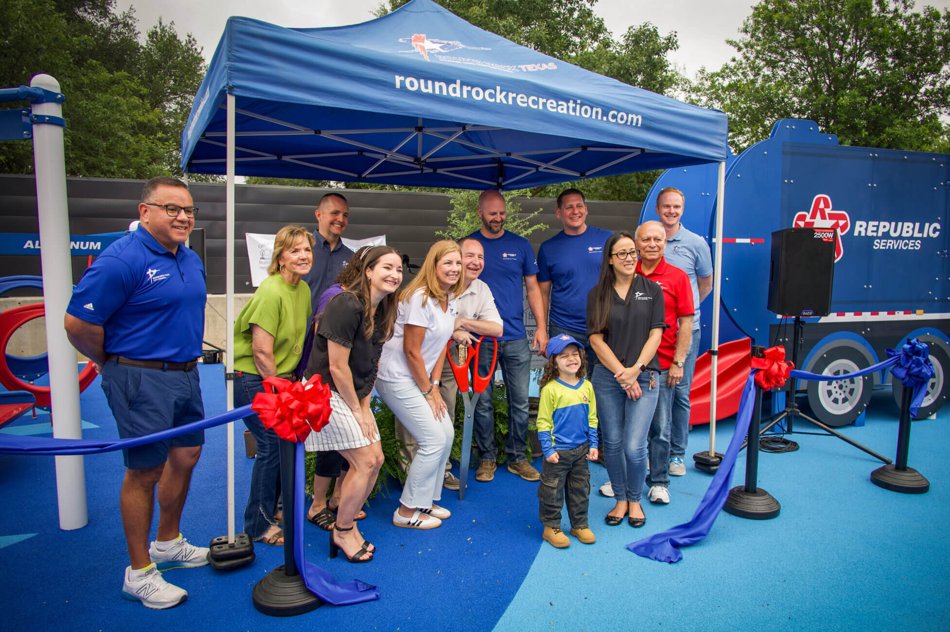 A group of people poses and smiles under a blue tent with “roundrockrecreation.com” printed on it, at an outdoor event. Blue and red decorations and a Republic Services truck are in the background.