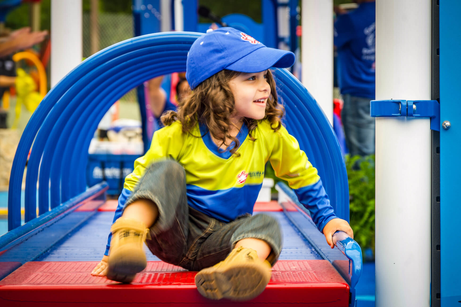 A young child in a blue cap and yellow shirt smiles while playing on blue and red playground equipment, sitting and leaning back with one knee up.