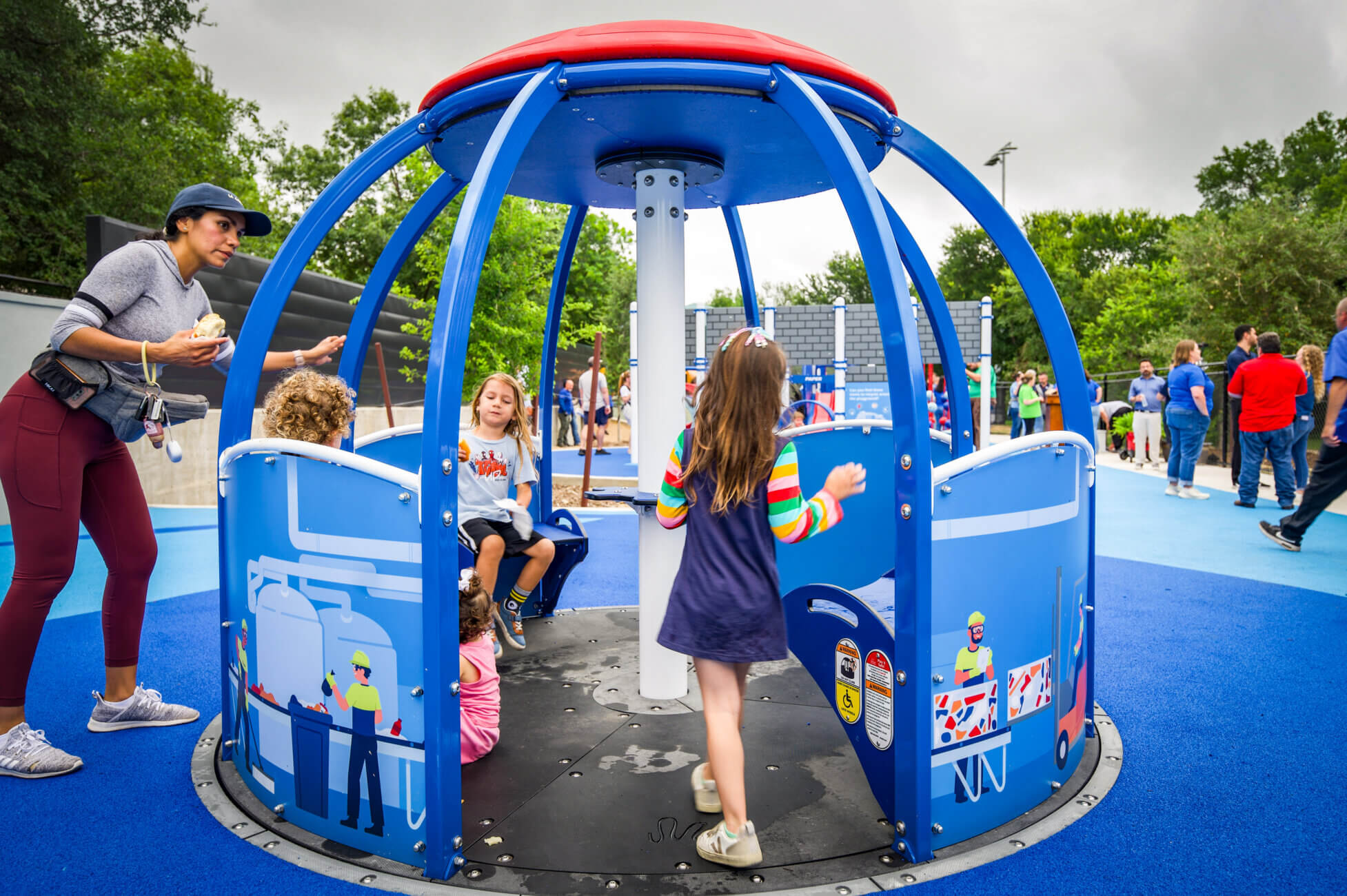 Children play inside a blue, round spinning playground structure, while an adult stands nearby. The playground has soft blue flooring, and other people are visible in the background under a cloudy sky.
