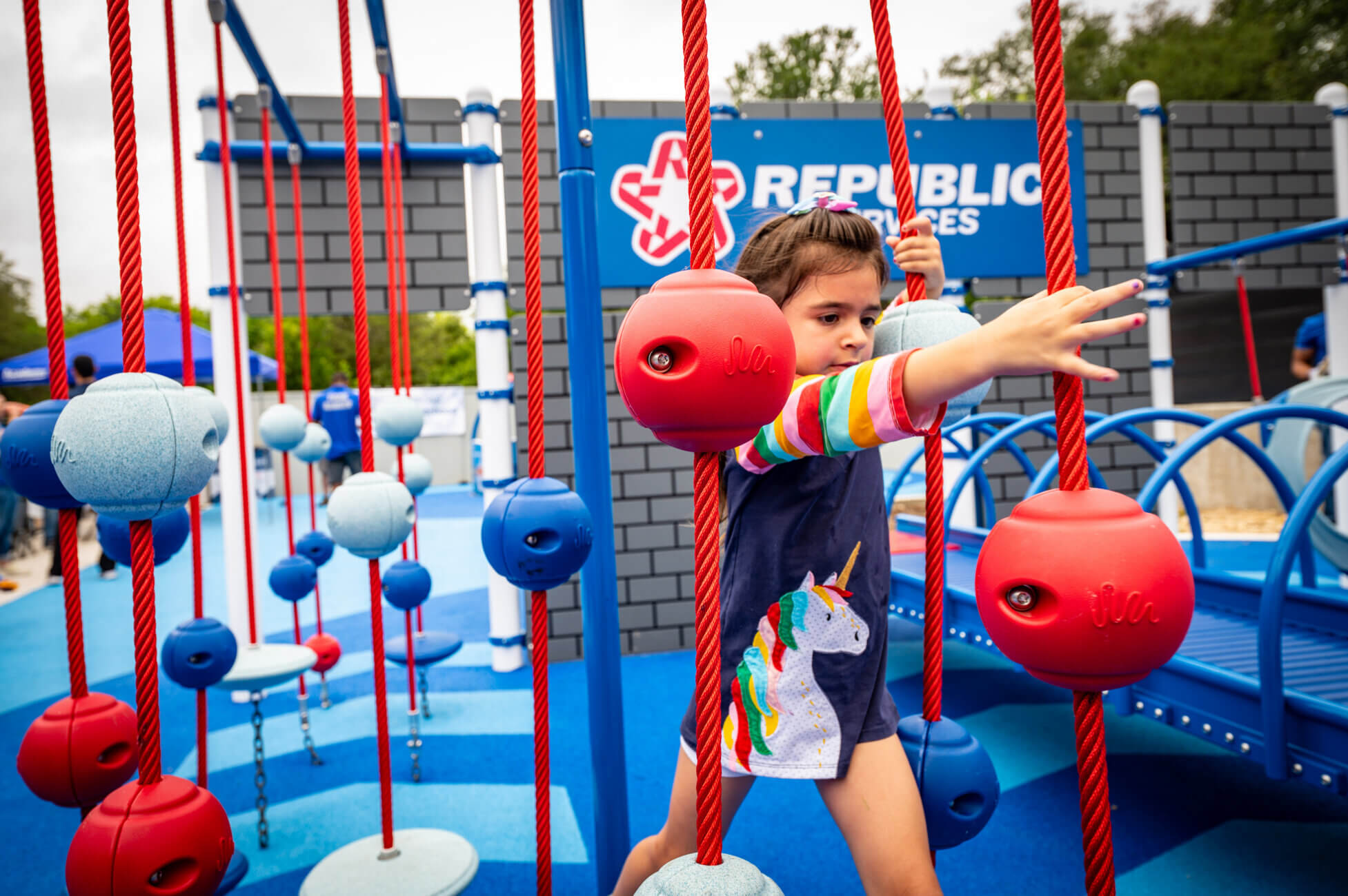 A young girl wearing a unicorn dress navigates a playground obstacle course with red and blue climbing balls and ropes. A blue play structure and Republic Services sign are visible in the background.