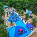 Aerial view of a vibrant blue playground filled with children and adults. The area features modern play structures, shaded seating, trees, and a large digital screen, surrounded by greenery.