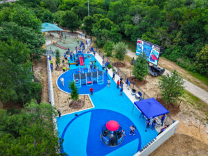 Aerial view of a vibrant blue playground filled with children and adults. The area features modern play structures, shaded seating, trees, and a large digital screen, surrounded by greenery.