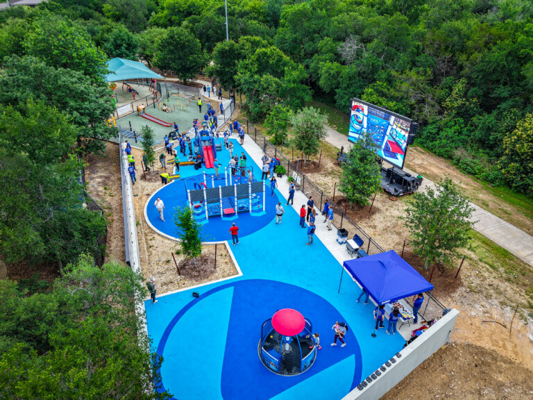 Aerial view of a vibrant blue playground filled with children and adults. The area features modern play structures, shaded seating, trees, and a large digital screen, surrounded by greenery.