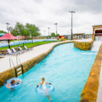 Two children float on inner tubes in a lazy river at a water park while an adult stands nearby on the pool deck. The sky is cloudy, and the area is surrounded by greenery and various park amenities.