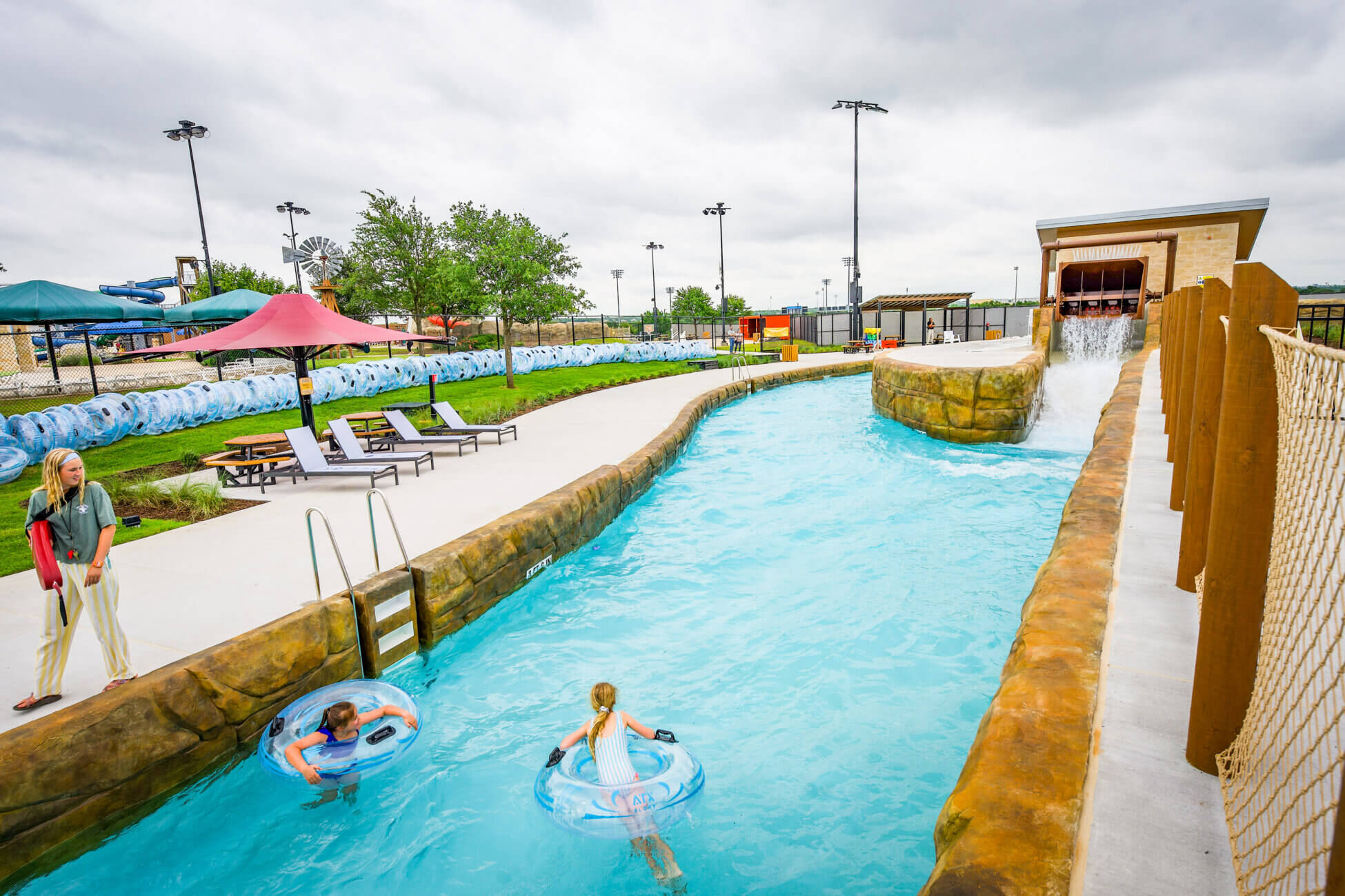 Two children float on inner tubes in a lazy river at a water park while an adult stands nearby on the pool deck. The sky is cloudy, and the area is surrounded by greenery and various park amenities.