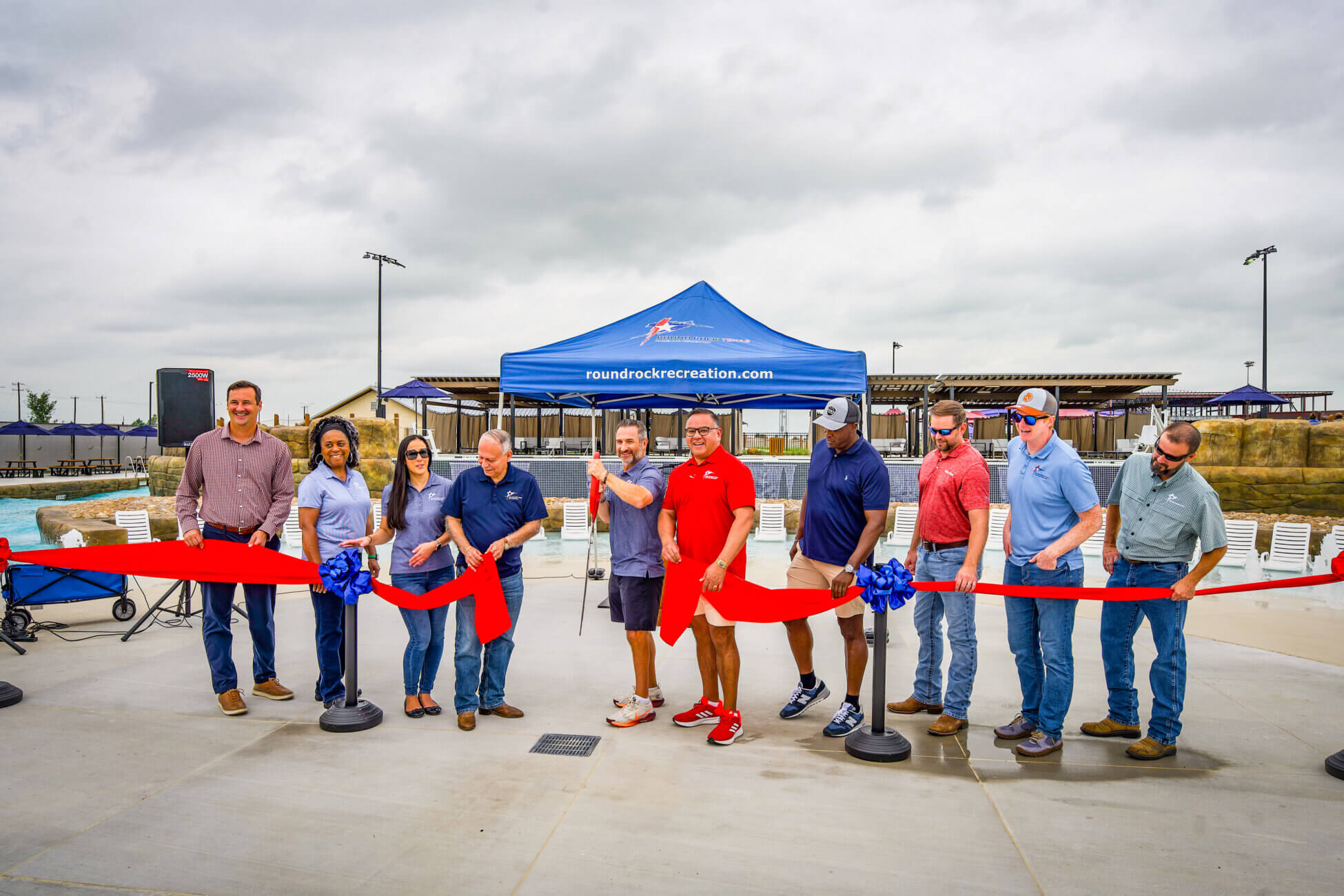 A group of people stand in front of a blue tent cutting a ceremonial red ribbon at an outdoor event, celebrating the opening of a recreation facility under cloudy skies.