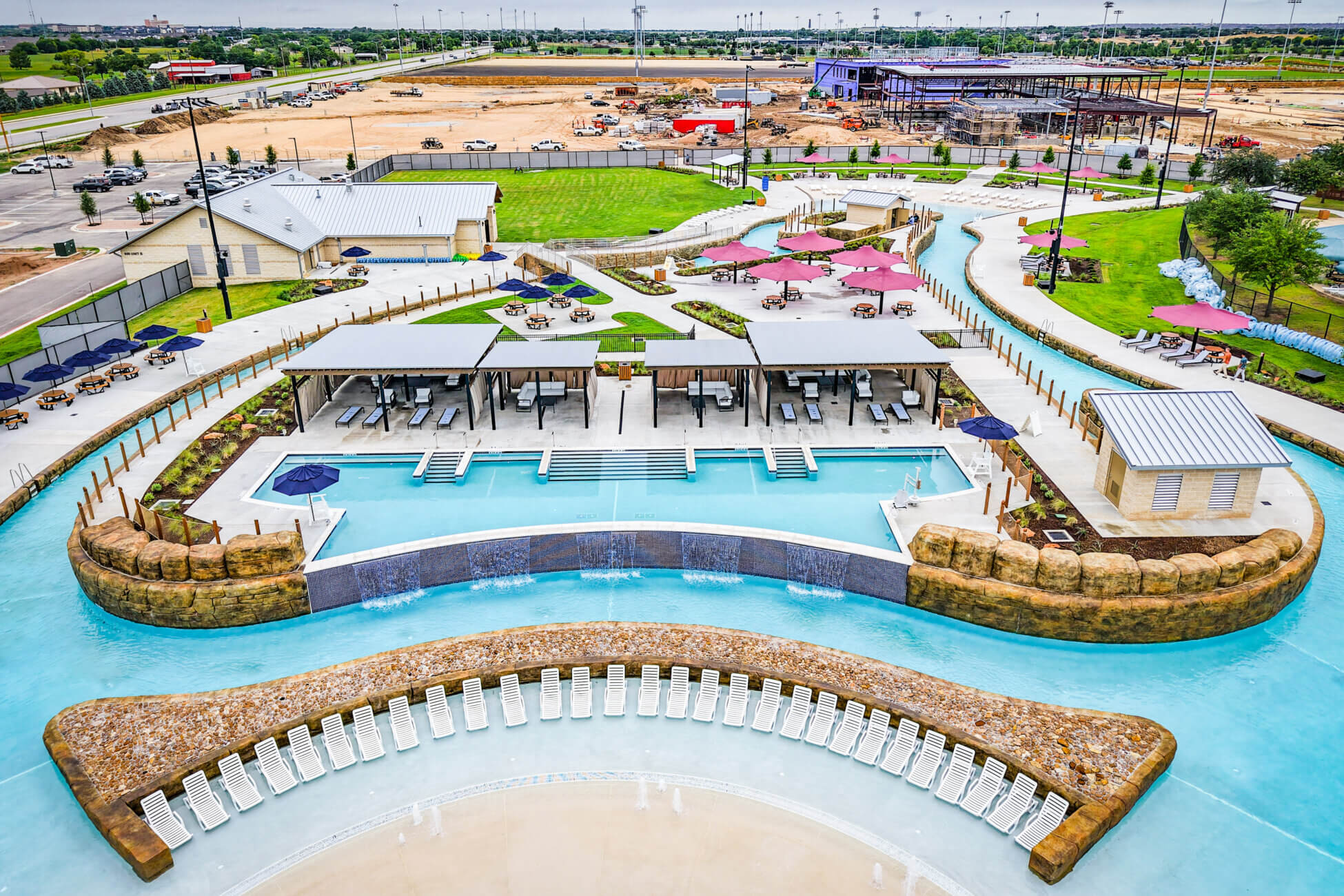 Aerial view of a modern water park featuring a lagoon-style pool with lounge chairs, cabanas, umbrellas, and a lazy river, surrounded by landscaped lawns and ongoing construction in the background.