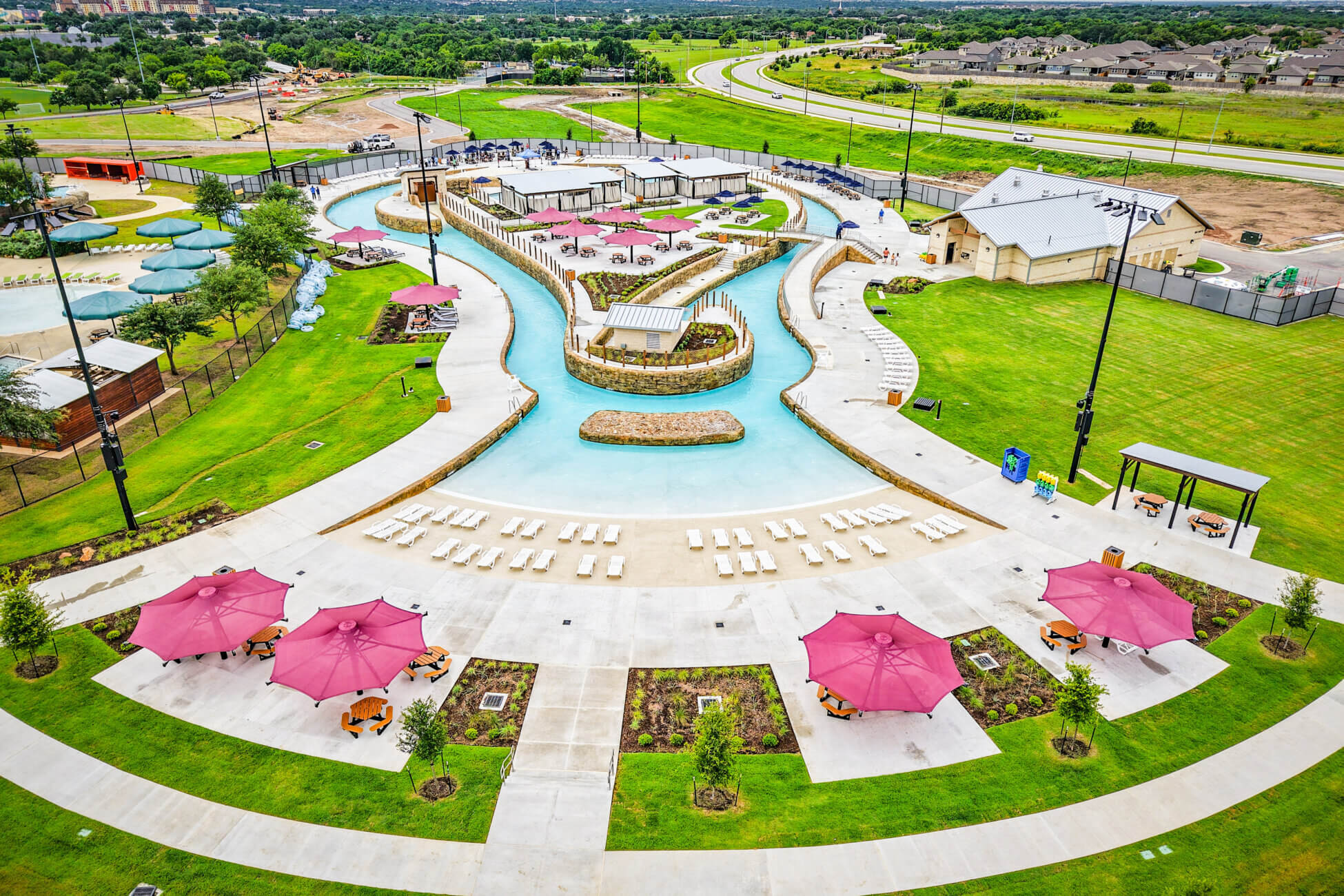 Aerial view of a landscaped outdoor pool area with a winding lazy river, lounge chairs, red umbrellas, shaded cabanas, green lawns, and nearby roads and rooftops in the background.