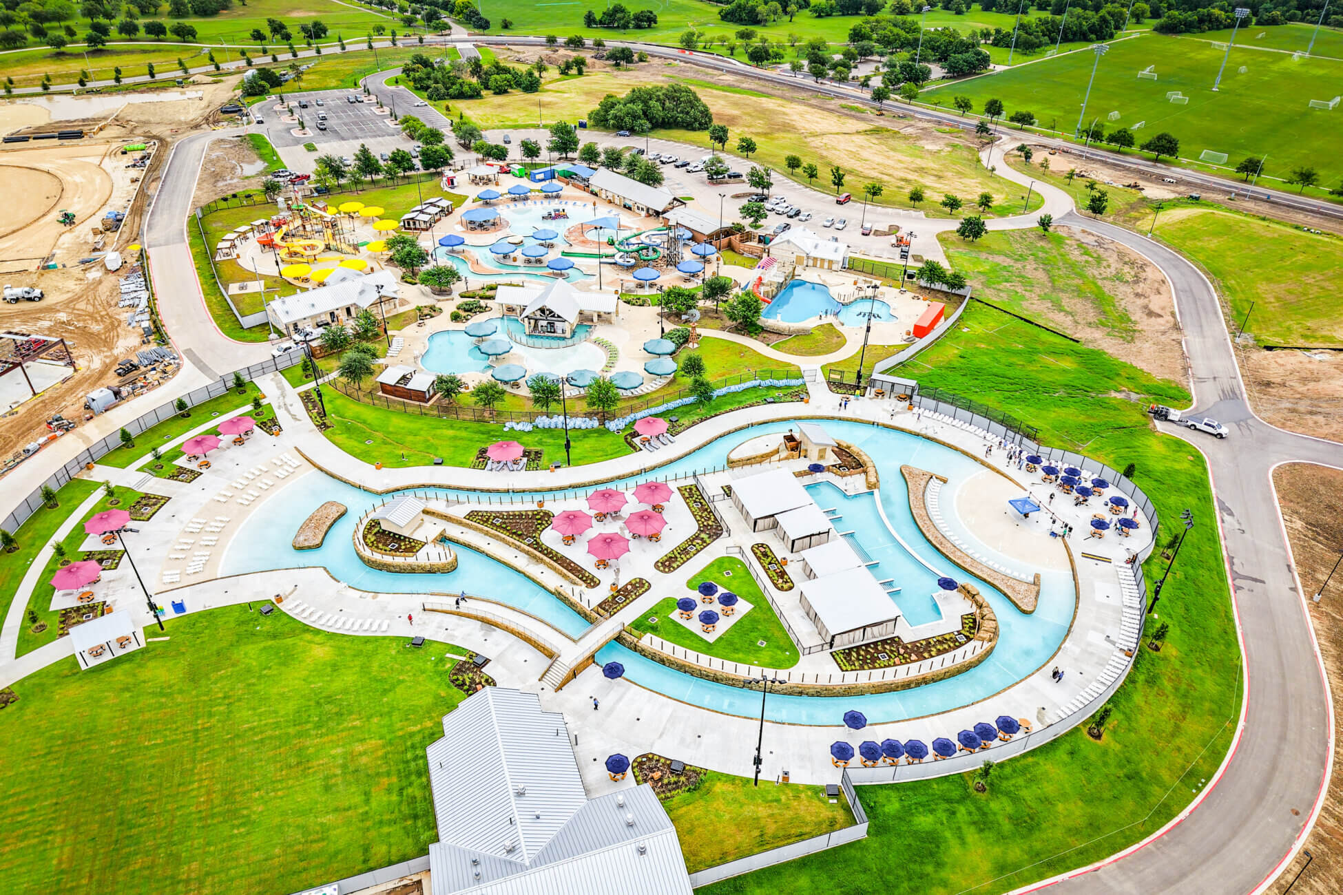 Aerial view of a water park with pools, water slides, a winding lazy river, lounge chairs, shaded umbrellas, and surrounding green lawns, with parking spaces and roads nearby.