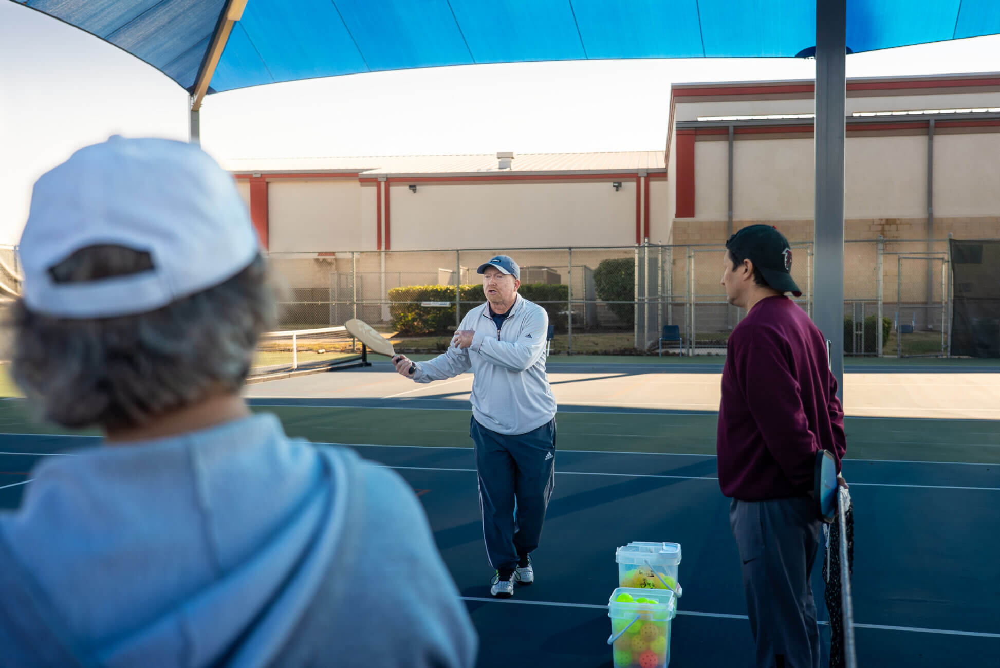 A coach holds a pickleball paddle and instructs two players on an outdoor court under a blue canopy, with a container of balls nearby.
