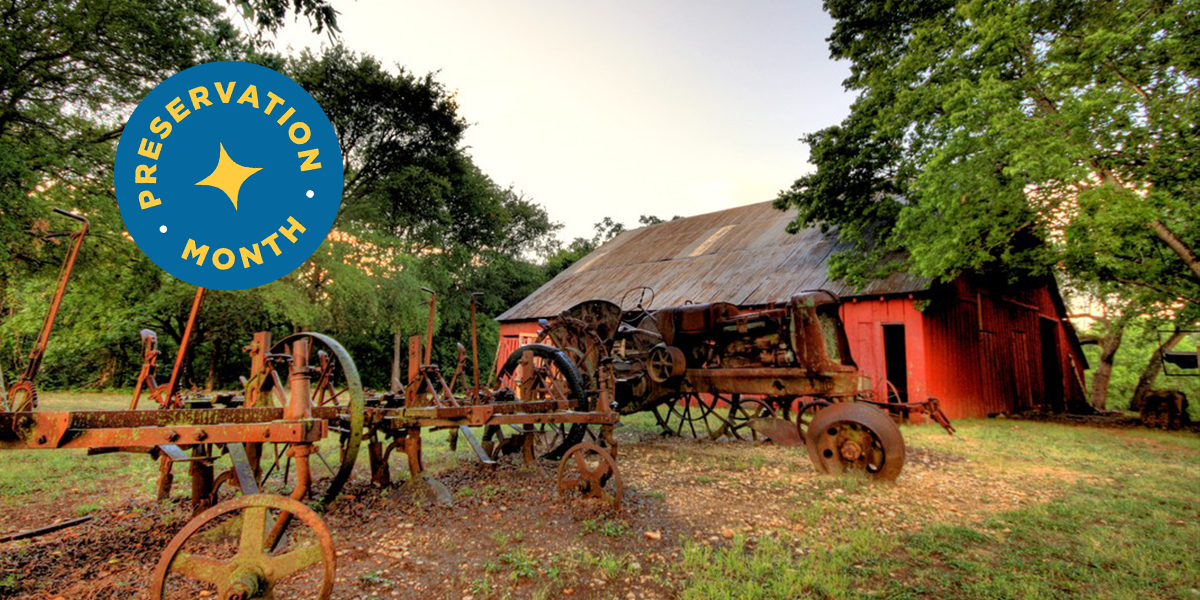 A rusty, vintage farm tractor and equipment sit near a weathered red barn among trees, with a blue Preservation Month badge in the top left corner.