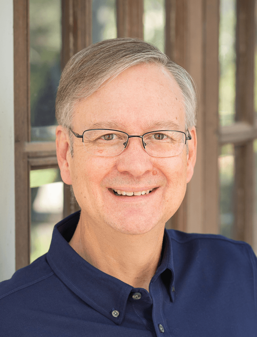 A middle-aged man with short gray hair and glasses smiles at the camera. He is wearing a navy blue collared shirt and standing in front of a window with wooden frames.