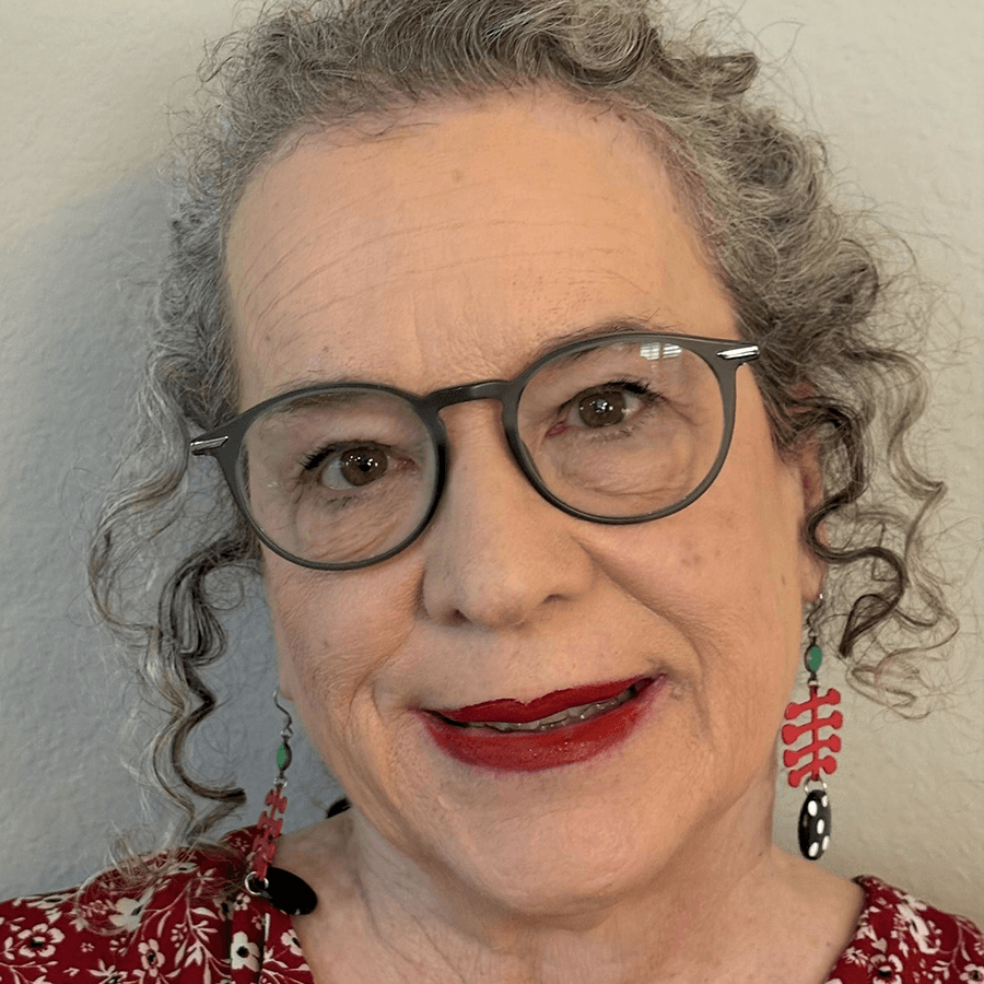 Smiling older person with curly gray hair, wearing round glasses, red lipstick, and red patterned earrings. They are dressed in a red and white patterned top, standing in front of a light-colored wall.