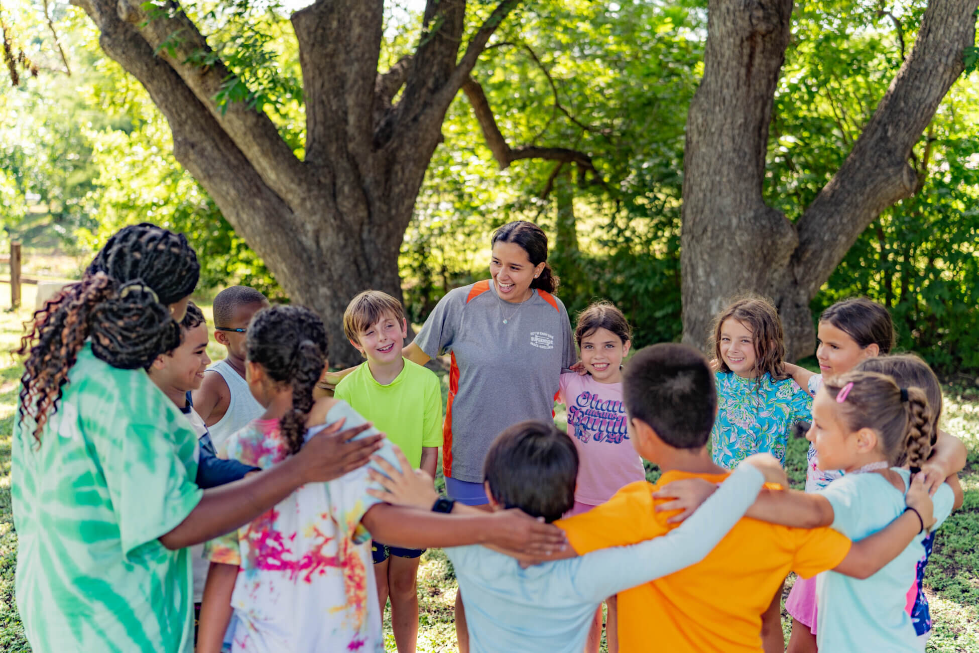 A group of children stands in a circle with their arms around each other, smiling, outdoors under large trees, while two adults supervise and join the group. The mood is cheerful and friendly.