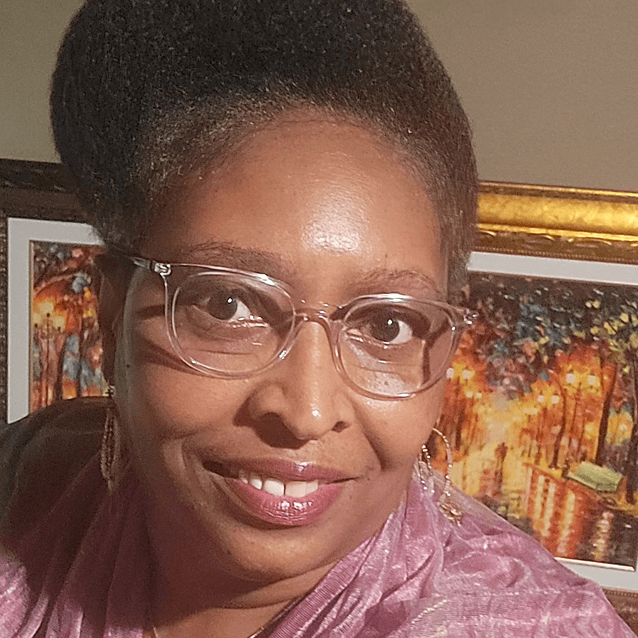 A woman with short, natural hair and clear glasses smiles at the camera. She wears a pink scarf and gold earrings. In the background, there is a colorful framed painting of a park scene with lights.