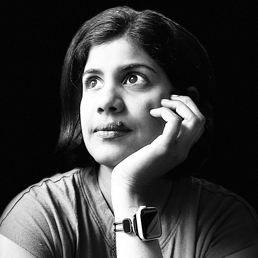 Black and white photo of a woman with short hair gazing upwards thoughtfully. She rests her chin on her hand and wears a smartwatch on her wrist. The background is dark, highlighting her face.
