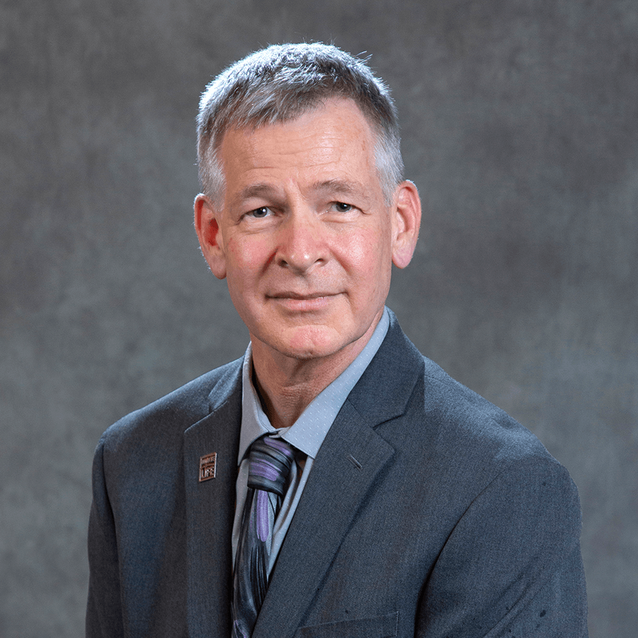 A middle-aged man with short gray hair wearing a gray suit, striped tie, and a pin on his lapel, sitting in front of a plain gray background and looking at the camera with a slight smile.