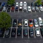 Aerial view of a full parking lot with rows of parked cars, including a noticeable bright green car among darker-colored vehicles, and a few small trees separating parking sections.