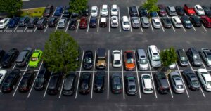 Aerial view of a full parking lot with rows of parked cars, including a noticeable bright green car among darker-colored vehicles, and a few small trees separating parking sections.
