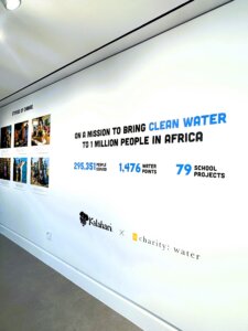 A white wall display notes a mission to bring clean water to 1 million people in Africa, with stats: 295,351 people served, 1,476 water points, 79 school projects. Logos for Kalahari and Charity: Water are at the bottom.