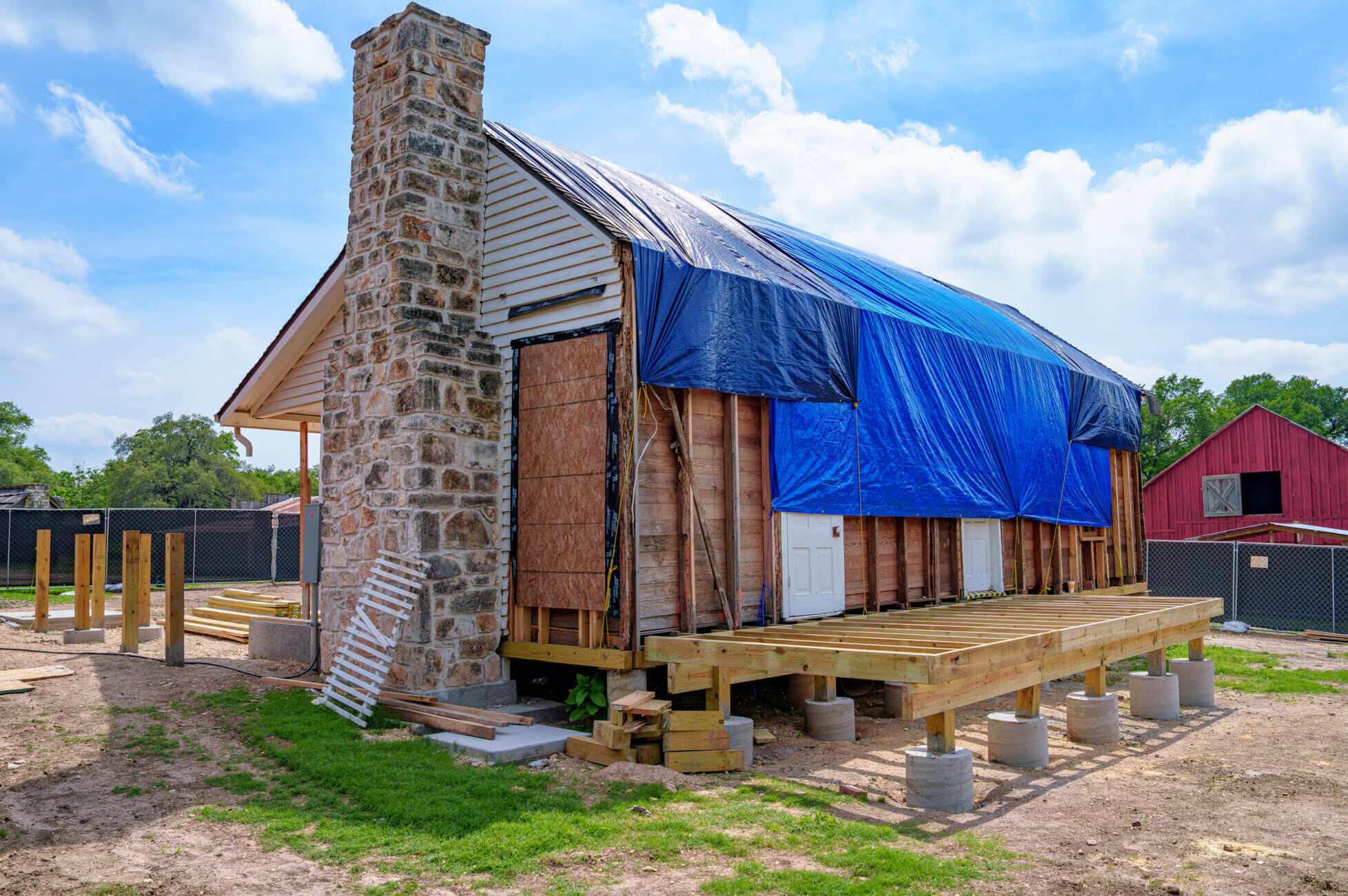 A small house under renovation, with its exterior covered by blue tarps, exposed wooden framing, a stone chimney, and a partially built porch on raised concrete piers. The sky is blue with scattered clouds.
