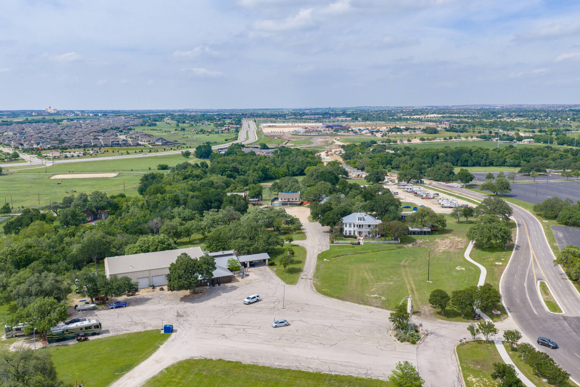 Aerial view of a suburban area with houses, trees, open grassy spaces, roads, and parking lots under a partly cloudy sky. Residential neighborhoods and fields stretch into the distance.