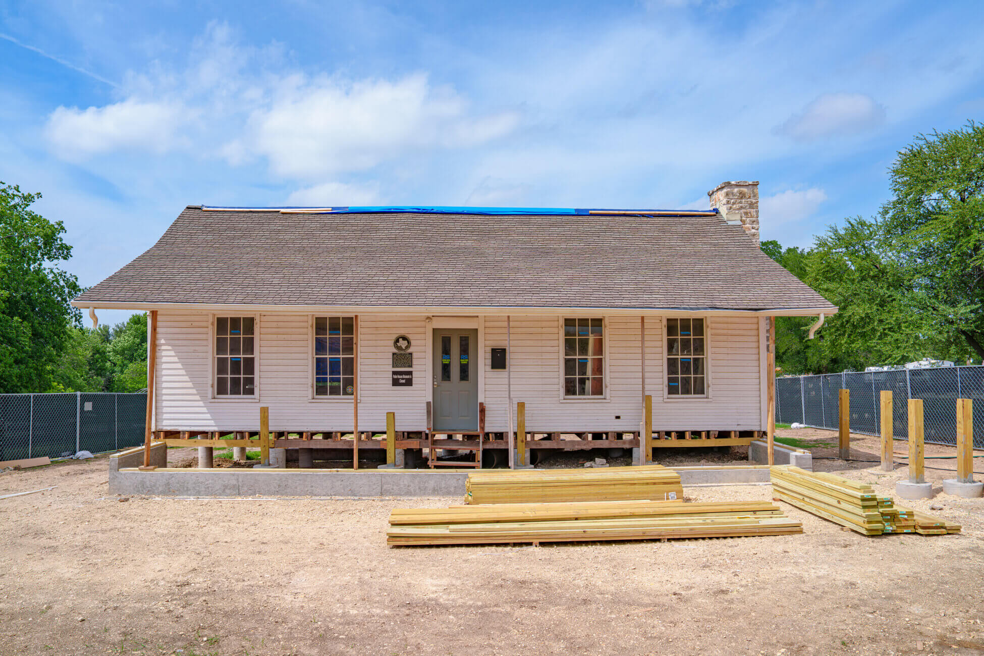 A single-story house under renovation with exposed foundation and unfinished front steps, surrounded by construction materials and fenced-off area, under a partly cloudy sky.