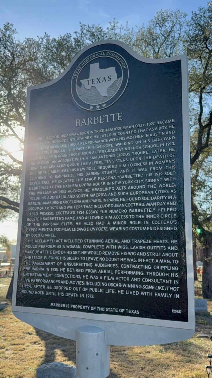 A historical marker titled Barbette stands in a cemetery, with sunlight shining through tree branches behind it. The plaque details the life and career of the aerialist and performer Barbette.