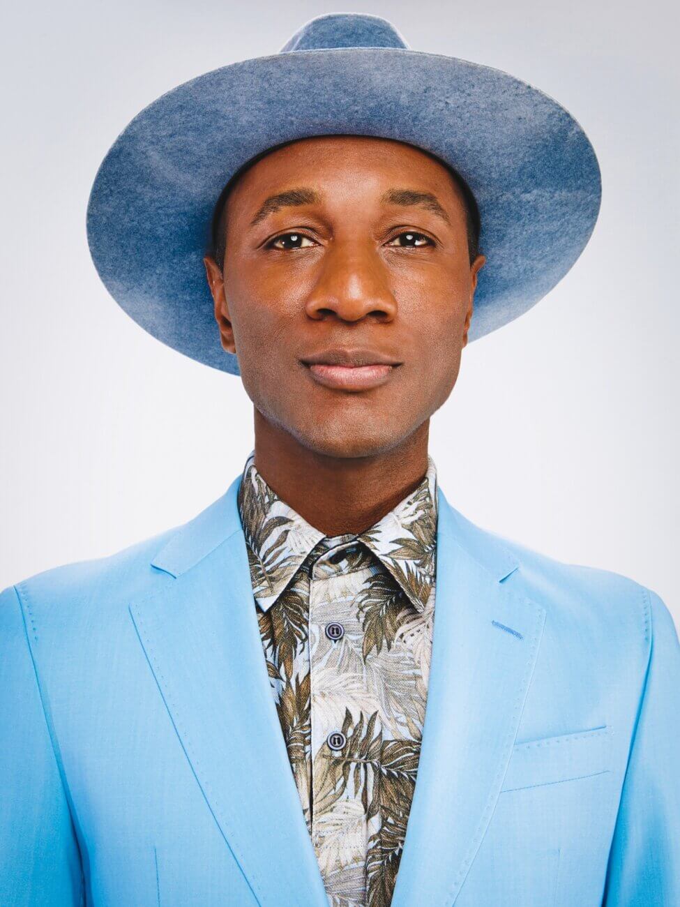 A person wearing a light blue suit jacket, a patterned shirt with leaves, and a matching wide-brimmed hat stands against a plain, light background, looking directly at the camera with a neutral expression.