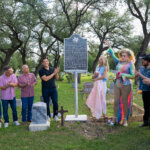 A group of people stands around a historical marker in a cemetery, applauding as a person in a colorful outfit and boots gestures toward the sign. Trees surround the scene and gravestones are visible in the foreground.