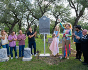 A group of people stands around a historical marker in a cemetery, applauding as a person in a colorful outfit and boots gestures toward the sign. Trees surround the scene and gravestones are visible in the foreground.
