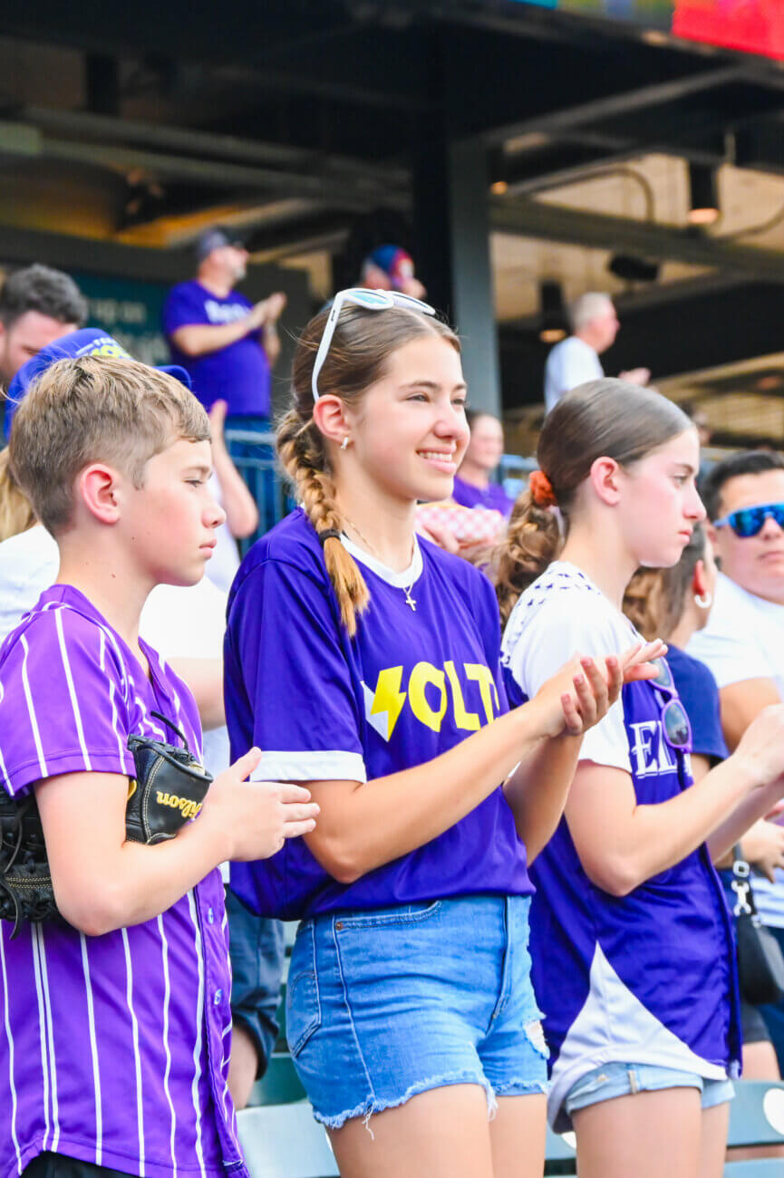 Three kids in purple shirts stand and clap at a sports event, with other fans in the background. One girl in the center smiles, wearing sunglasses on her head, while the others look toward the field.