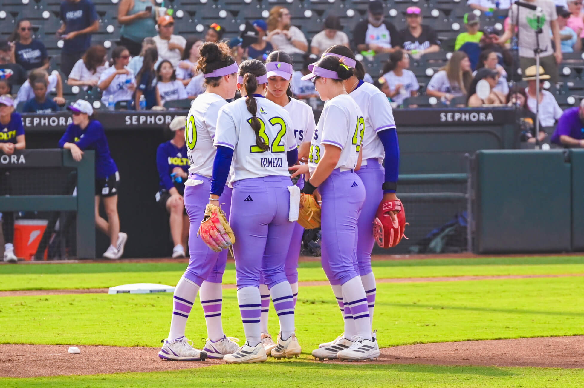 Four softball players in purple and white uniforms stand together in a huddle on a baseball field, with a crowd of spectators visible in the stadium seats behind them.