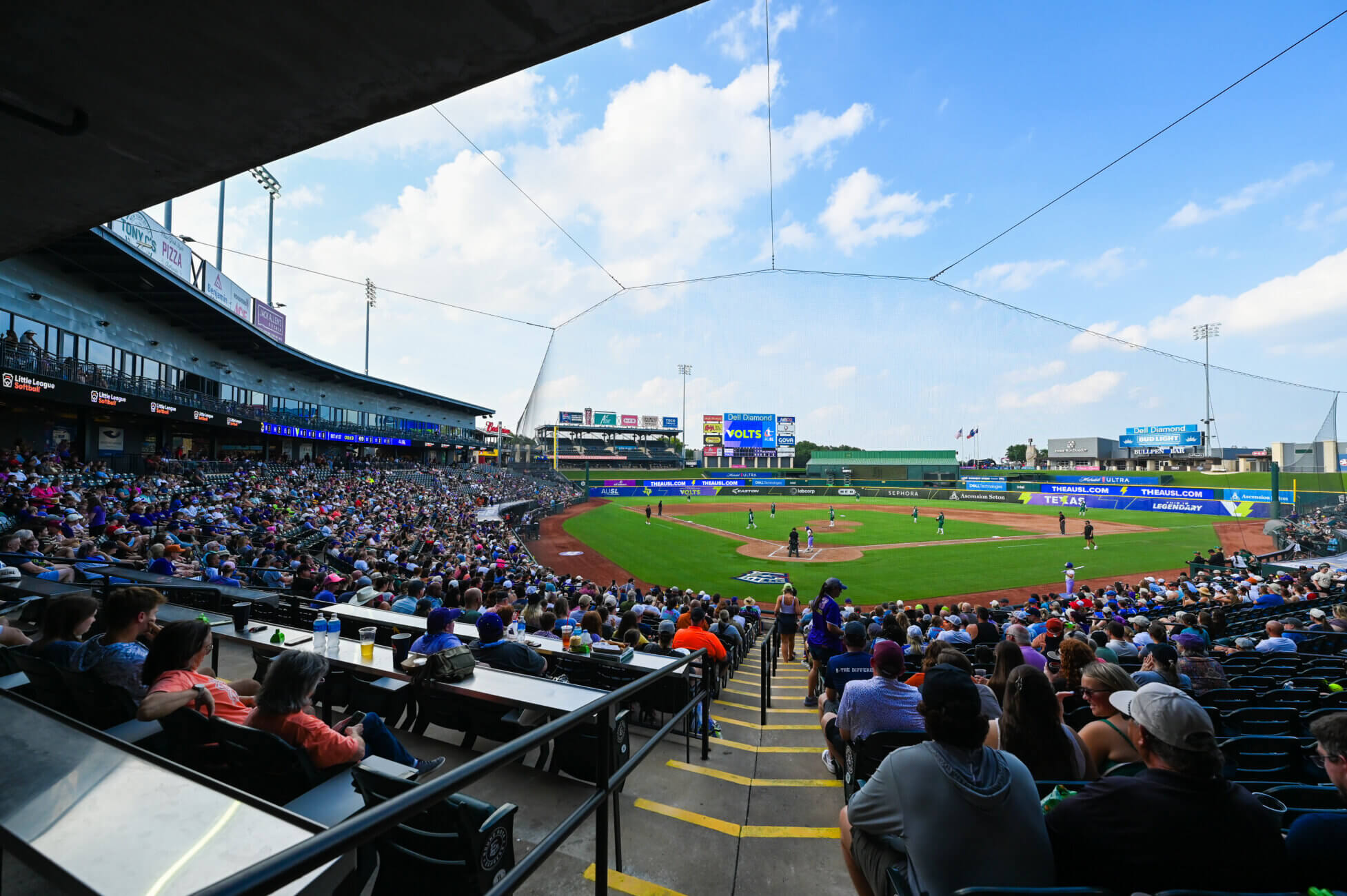 A large crowd fills the stands at a baseball stadium on a sunny day, watching a game in progress on the field. Fans sit in both shaded and open areas, with the diamond and outfield clearly visible.