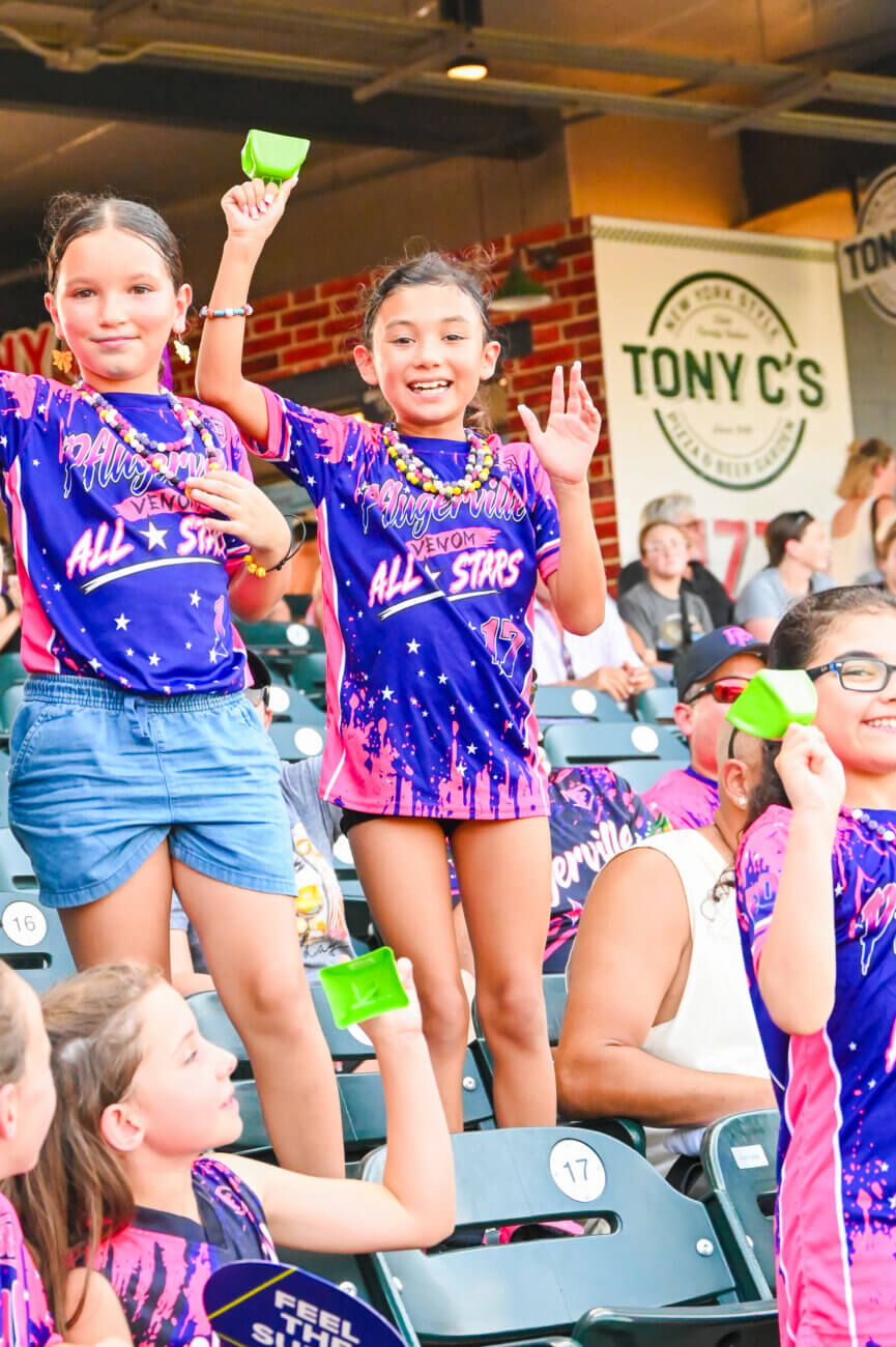 A group of smiling young girls in matching purple and pink All Stars shirts cheer and wave green cups while standing on stadium seats at a sports event. Other spectators and signage are visible in the background.