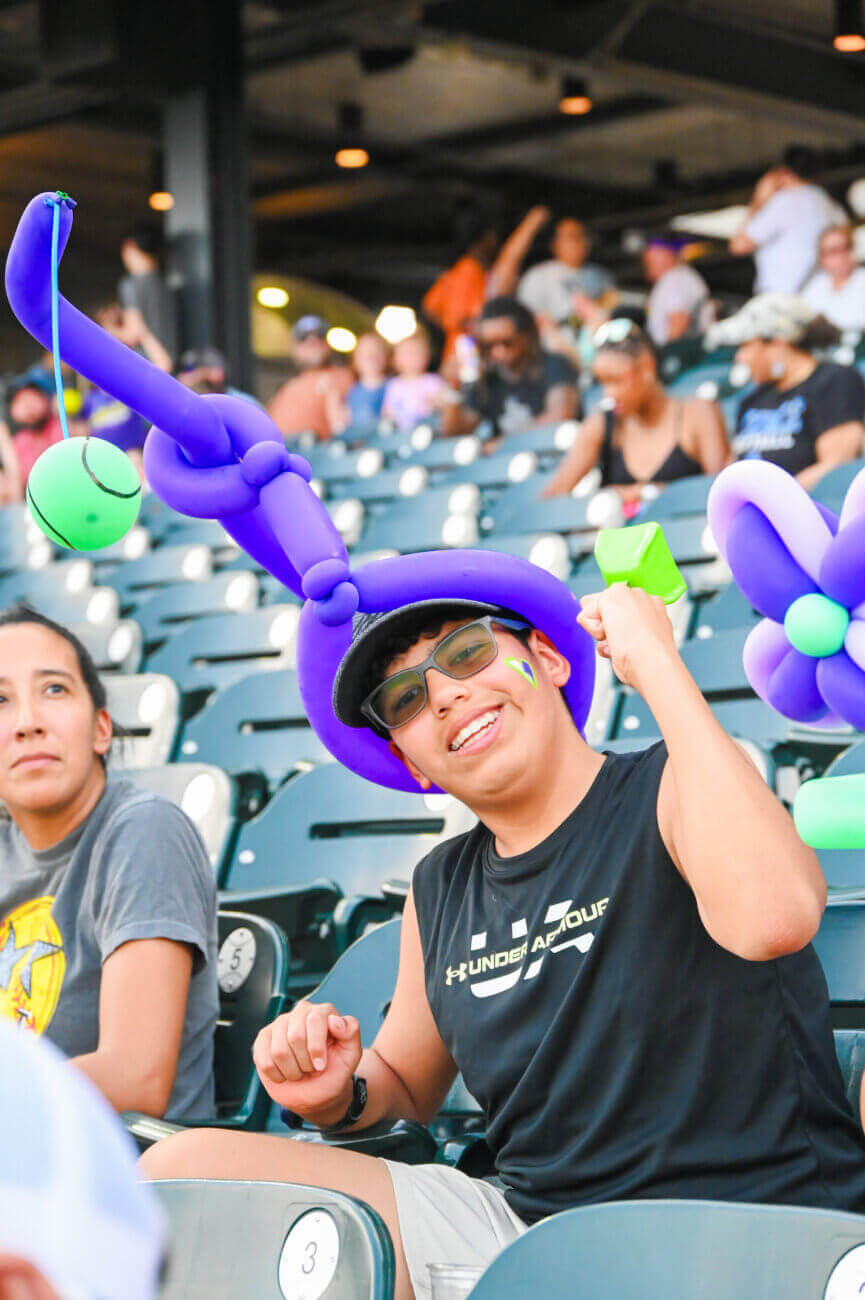 A smiling person wearing sunglasses and a purple balloon hat with green decorations sits in stadium seats, holding a green object. Other spectators and empty seats are visible in the background.