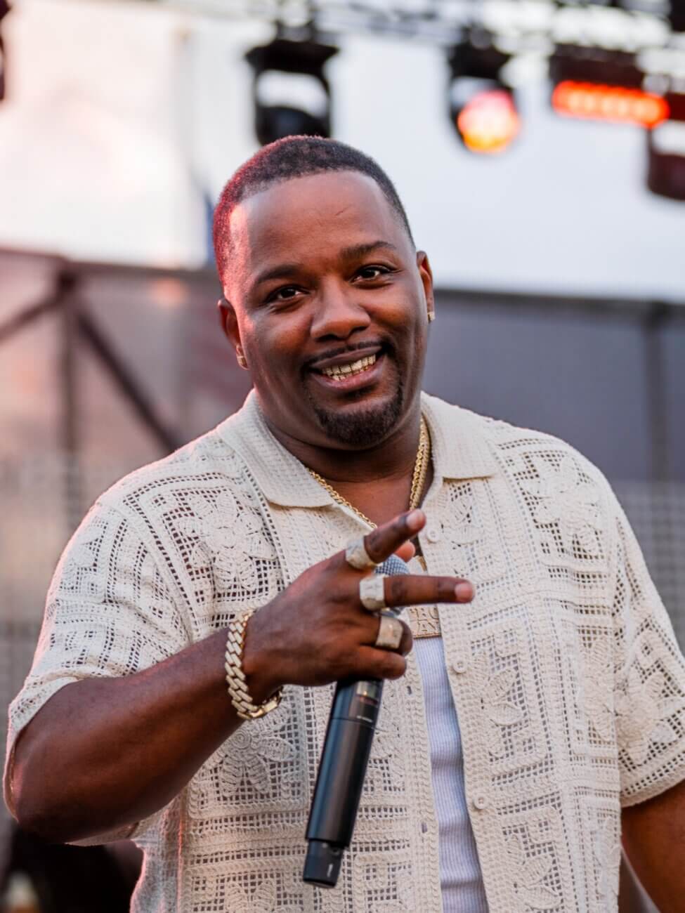 A man wearing a patterned short-sleeve shirt smiles while holding a microphone and making a peace sign with his hand. He wears jewelry and stands in front of stage lights and equipment.