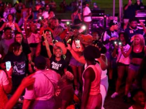 A lively crowd enjoys an outdoor event at night, with many people smiling, dancing, and holding up phones with flashlights. The scene is brightly lit with pink and purple stage lights.